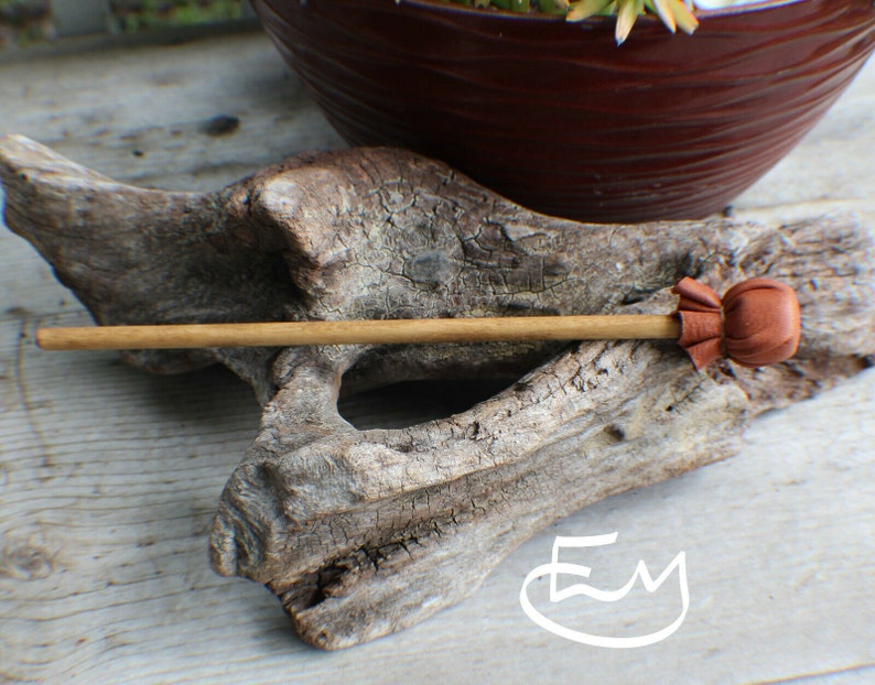 May include: A wooden hair stick with a brown leather accent. The hair accessory is set against a weathered wood background, with a brown pot and greenery in the background. The hair stick is a natural color.