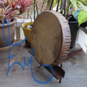 May include: A round, tan-colored hand drum with a textured, natural skin head and a wooden frame. The drum is positioned upright on a dark brown, angled metal stand, displaying a smooth, circular surface with subtle variations in tone.