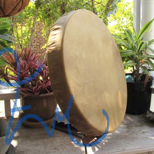 May include: A round, tan-colored frame drum with a smooth, taut skin head stretched over a circular wooden frame. The drum is positioned upright on a dark metal stand, displaying a uniform, light brown surface texture across the drumhead.