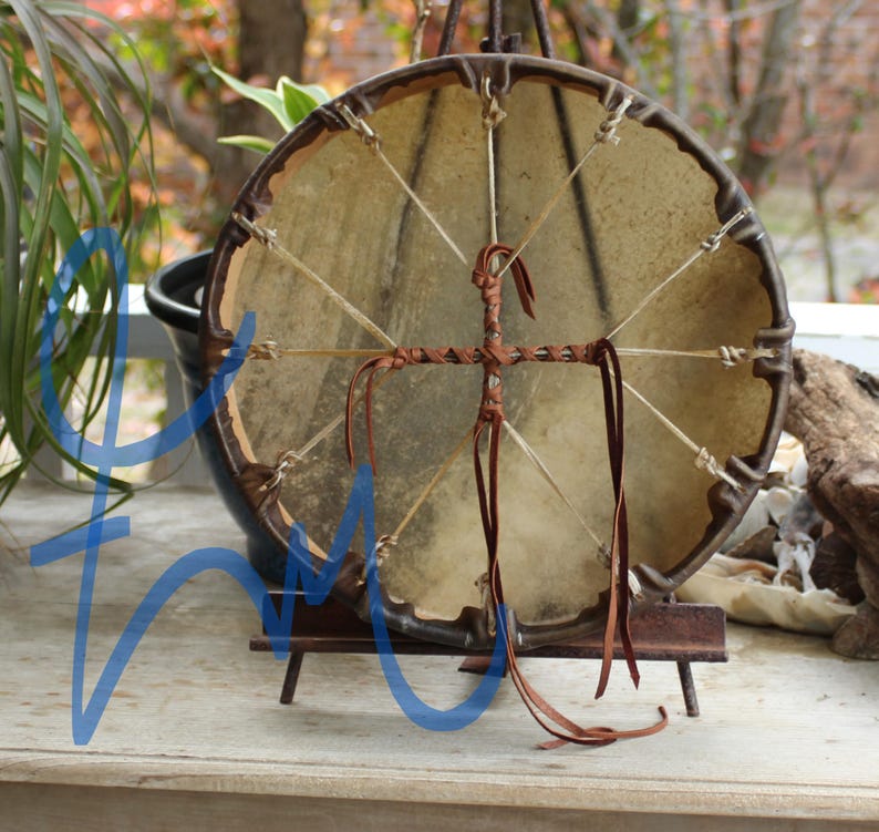 May include: A round, handcrafted drum with a natural hide drumhead. The drum has a dark brown wooden frame and is decorated with leather accents. The drum is displayed on a small wooden stand, with a blurred background of foliage.