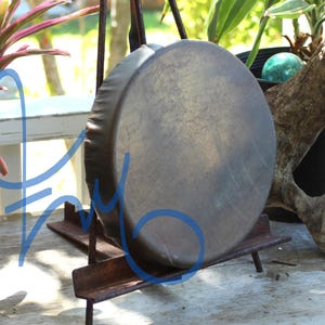 May include: A large, round, bronze-colored gong is mounted on a rusty metal stand. The gong has a slightly textured surface and is positioned outdoors. The background includes greenery and a piece of driftwood.