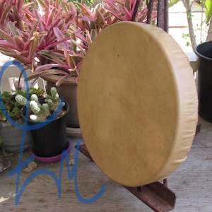May include: A circular frame drum with a smooth, light tan, opaque drumhead surface. The drum is positioned upright on a dark metal stand, displaying a uniform, solid color across its front face and a wrapped edge around the circular frame.