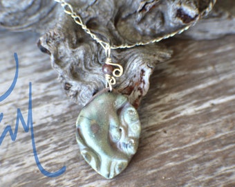 May include: A handcrafted pendant necklace featuring a unique, leaf-shaped pendant with a textured, iridescent finish in shades of green and brown. The pendant is suspended from a delicate gold-colored chain, with a small red bead accent. The necklace is displayed on a wooden surface.