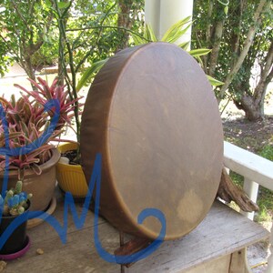 May include: A round, hand-held drum with a smooth, tan-colored animal hide head stretched over a circular wooden frame. The drum surface displays natural variations in color and texture, featuring a consistent, taut, and circular design.