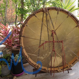 May include: A circular frame drum with a tan rawhide head stretched over a wooden hoop. The drum features a brown leather lacing system that secures the head to the frame and includes a central cross-shaped leather handle on the back.