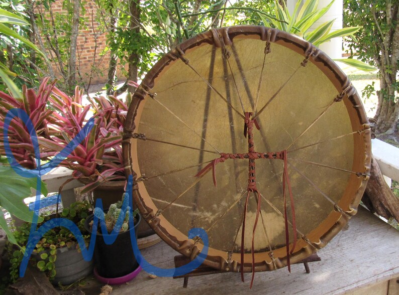 May include: A circular frame drum with a tan rawhide head stretched over a wooden hoop. The drum features a brown leather lacing system that secures the head to the frame and includes a central cross-shaped leather handle on the back.