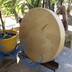 May include: A round, beige, hand-held drum with a smooth, taut skin surface stretched over a circular frame. The drum is positioned upright on a dark metal stand, displaying a natural, light tan color with subtle variations in the texture of the skin.