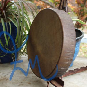 May include: A large, round, brown drum with a textured surface. The drum is suspended on a metal stand. The background includes potted plants and a blurred outdoor setting.