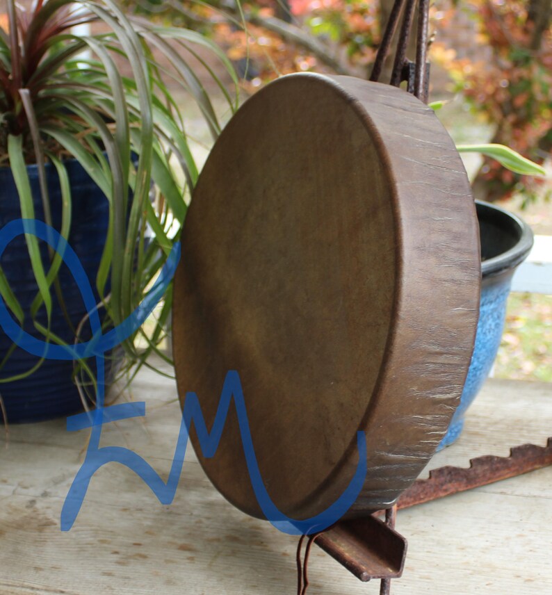 May include: A large, round, brown drum with a textured surface. The drum is suspended on a metal stand. The background includes potted plants and a blurred outdoor setting.