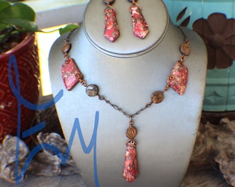 May include: A necklace and earring set displayed on a mannequin. The jewelry features red and brown patterned stones, copper wire, and a chain. The earrings are long, and the necklace has a pendant. The set is displayed on a gray mannequin.