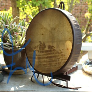 Puede incluir: Un tambor grande y redondo con un parche de piel natural y un marco marr&oacute;n oscuro. El tambor est&aacute; sostenido por un soporte de metal oscuro. El parche del tambor tiene un aspecto desgastado y envejecido. Un asa est&aacute; unida a la parte superior del tambor.