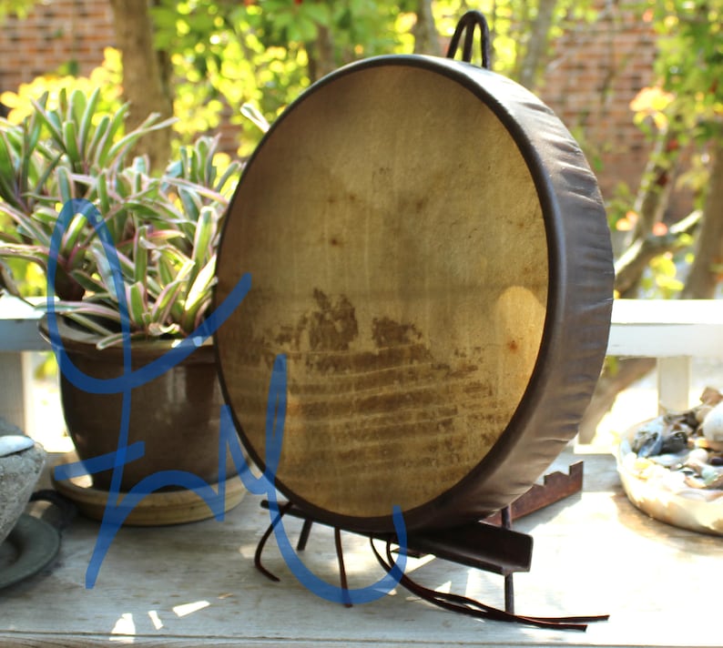 Puede incluir: Un tambor grande y redondo con un parche de piel natural y un marco marr&oacute;n oscuro. El tambor est&aacute; sostenido por un soporte de metal oscuro. El parche del tambor tiene un aspecto desgastado y envejecido. Un asa est&aacute; unida a la parte superior del tambor.