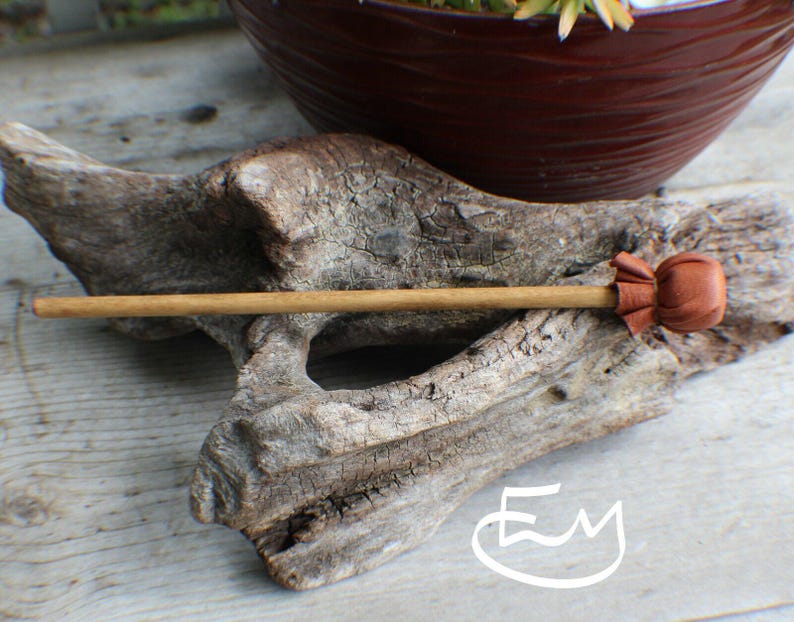 May include: A straight wooden hair stick featuring a smooth, light brown cylindrical shaft and a decorative, rounded brown leather accent piece at one end. The leather topper is gathered and secured to the wooden stick.