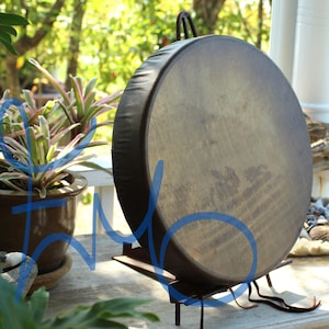 Puede incluir: Un tambor grande y redondo con una superficie de cuero marr&oacute;n oscuro y un borde negro. El tambor est&aacute; montado sobre un soporte de metal negro. El fondo incluye plantas en macetas y un entorno exterior borroso.