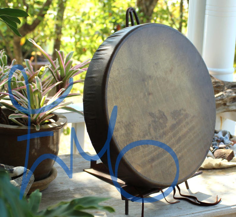 Puede incluir: Un tambor grande y redondo con una superficie de cuero marr&oacute;n oscuro y un borde negro. El tambor est&aacute; montado sobre un soporte de metal negro. El fondo incluye plantas en macetas y un entorno exterior borroso.