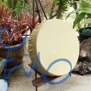 May include: A circular frame drum with a smooth, light beige skin head stretched tightly over a wooden rim. The drum is mounted on a dark, rusted metal stand, displaying a clean, solid surface without any visible markings or decorative patterns.