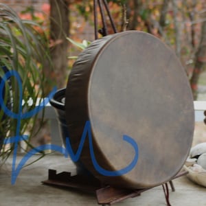 May include: A round, brown drum with a dark rim and a natural leather surface. The drum is supported by a dark metal stand. The background includes greenery and a glimpse of an outdoor setting.