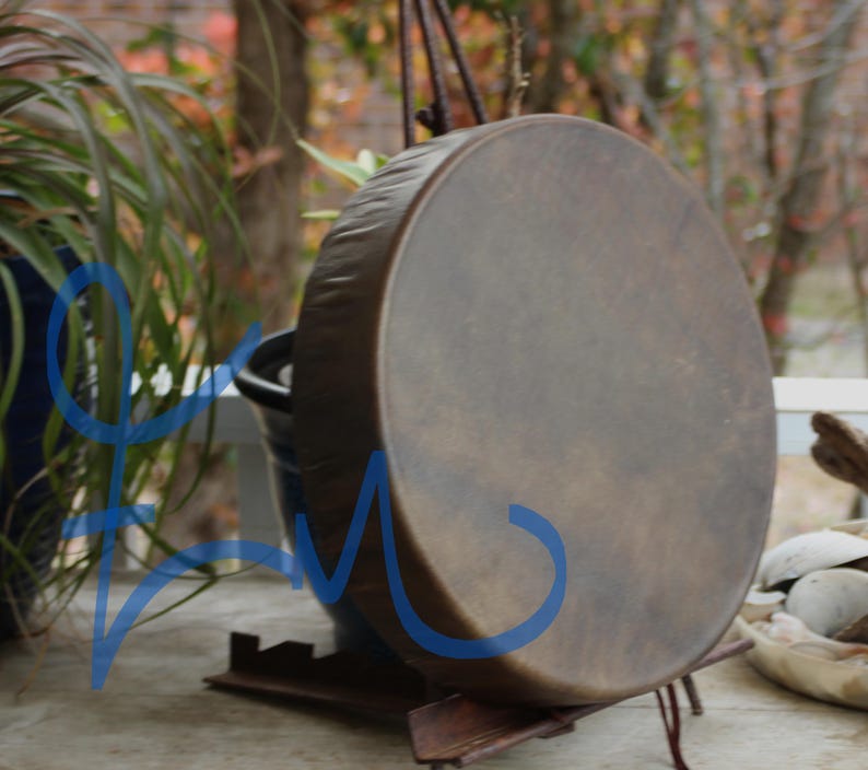 May include: A round, brown drum with a dark rim and a natural leather surface. The drum is supported by a dark metal stand. The background includes greenery and a glimpse of an outdoor setting.