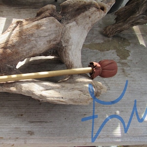 May include: A light-colored wooden stick featuring a rounded, reddish-brown leather topper secured with a small leather tie. The object is positioned horizontally across a piece of weathered, light-gray driftwood with a textured surface.