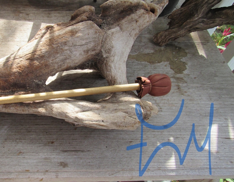 May include: A light-colored wooden stick featuring a rounded, reddish-brown leather topper secured with a small leather tie. The object is positioned horizontally across a piece of weathered, light-gray driftwood with a textured surface.