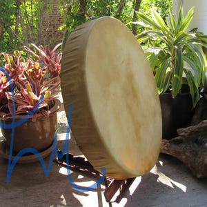 May include: A round frame drum with a light tan, natural skin head stretched over a wooden frame. The drum is positioned upright on a dark wooden stand, showcasing a smooth, circular surface with subtle variations in color and texture.