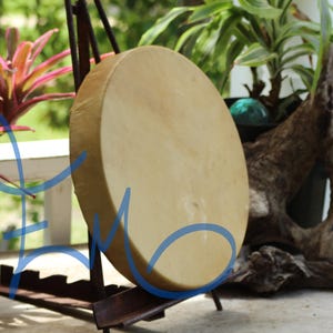 May include: A large, round drum with a light tan surface is displayed on a dark metal stand. The drum is the focal point, with a natural, organic aesthetic. The background includes greenery and a weathered wooden element.
