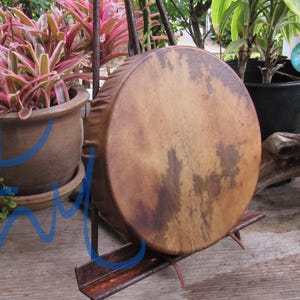 May include: A circular frame drum with a natural, mottled tan and brown animal hide head. The drum is mounted on a dark, angled metal stand that supports the base and back, displaying the textured surface of the drum skin clearly.