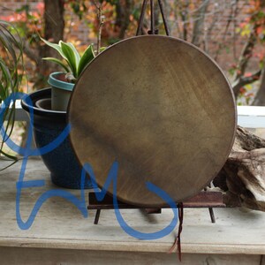 May include: A round, brown drum with a natural hide surface, suspended from a metal stand. The drum is displayed outdoors, with plants and a wooden surface in the background. The drum's color is a warm, earthy tone.