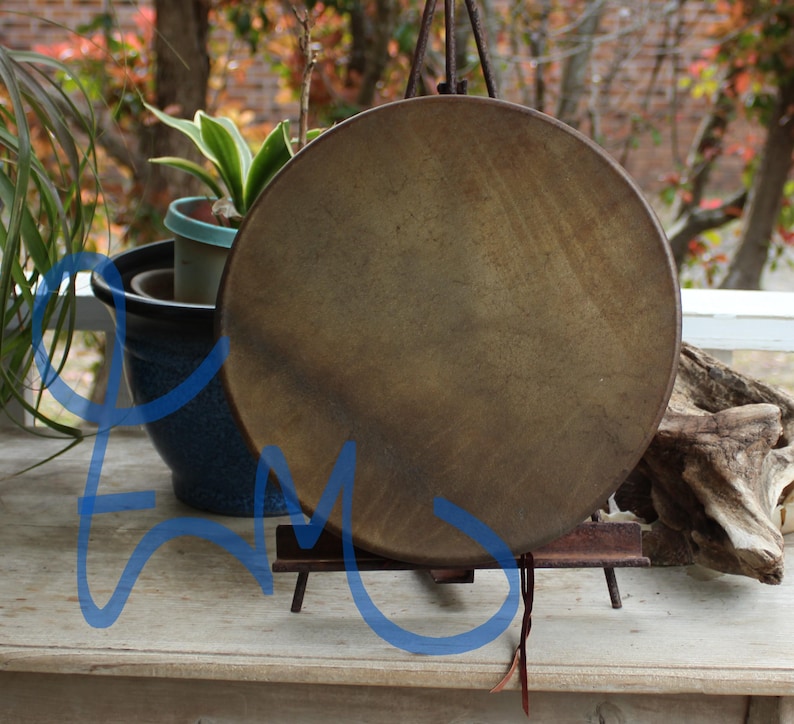 May include: A round, brown drum with a natural hide surface, suspended from a metal stand. The drum is displayed outdoors, with plants and a wooden surface in the background. The drum's color is a warm, earthy tone.