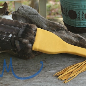 May include: A handcrafted leather item with a yellow handle and fringe, attached to a section of dark brown feathers. The item rests on a weathered wooden surface, with a turquoise pot in the background. The item appears to be a decorative piece.