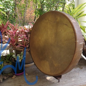 May include: A round frame drum with a tan, textured animal hide head stretched over a dark brown wooden circular frame. The drum is positioned upright on a small metal stand, displaying a smooth, taut surface with natural variations in color.