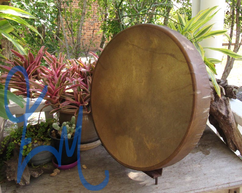 May include: A round frame drum with a tan, textured animal hide head stretched over a dark brown wooden circular frame. The drum is positioned upright on a small metal stand, displaying a smooth, taut surface with natural variations in color.