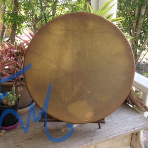May include: A circular frame drum with a smooth, tan-colored skin head stretched across a wooden rim. The drum is positioned upright on a small wooden stand, displaying a uniform, matte surface texture with subtle variations in the natural tan tone.