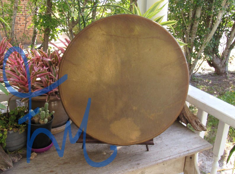 May include: A circular frame drum with a smooth, tan-colored skin head stretched across a wooden rim. The drum is positioned upright on a small wooden stand, displaying a uniform, matte surface texture with subtle variations in the natural tan tone.