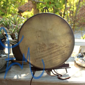 Puede incluir: Un gran tambor redondo de estilo antiguo con una superficie marr&oacute;n dorada desgastada. El tambor est&aacute; montado sobre un soporte de metal oscuro con un asa en la parte superior. El fondo incluye vegetaci&oacute;n y una superficie de madera.
