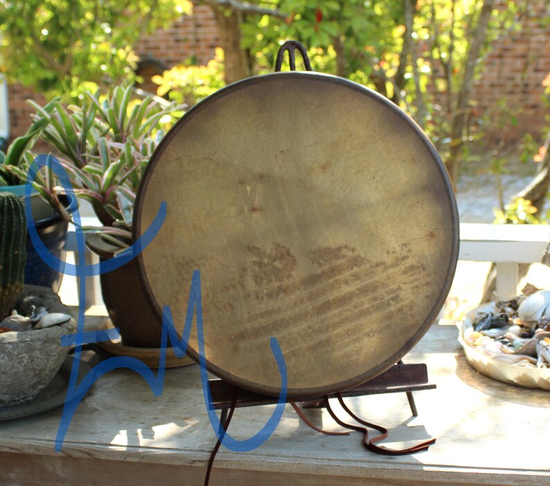 Puede incluir: Un gran tambor redondo de estilo antiguo con una superficie marr&oacute;n dorada desgastada. El tambor est&aacute; montado sobre un soporte de metal oscuro con un asa en la parte superior. El fondo incluye vegetaci&oacute;n y una superficie de madera.