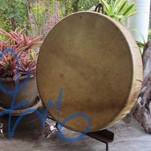 May include: A round, hand-held drum featuring a smooth, light tan animal hide head stretched tightly over a circular wooden frame. The drum is positioned upright on a dark metal stand, displaying a natural, mottled texture on the surface.