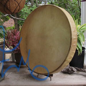 May include: A round frame drum with a light tan, smooth animal hide head stretched tightly over a wooden rim. The drum is positioned upright on a small metal stand, displaying a uniform, matte surface texture across the circular drumhead.