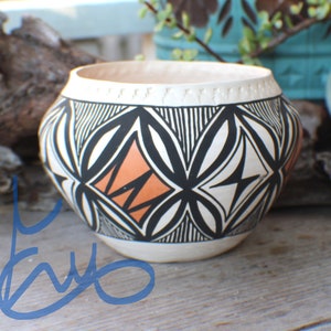 May include: A handcrafted ceramic bowl with a cream-colored interior and base. The exterior features a geometric design in black, white, and orange. The rim has a decorative pattern. This decorative bowl is perfect for home decor.