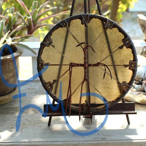 Puede incluir: Un tambor redondo, hecho a mano, con un parche de color beige claro y un marco de madera marr&oacute;n oscuro. El tambor est&aacute; sostenido por un peque&ntilde;o caballete marr&oacute;n oscuro. El dise&ntilde;o incluye un elemento en forma de cruz en el centro y detalles de cuero.