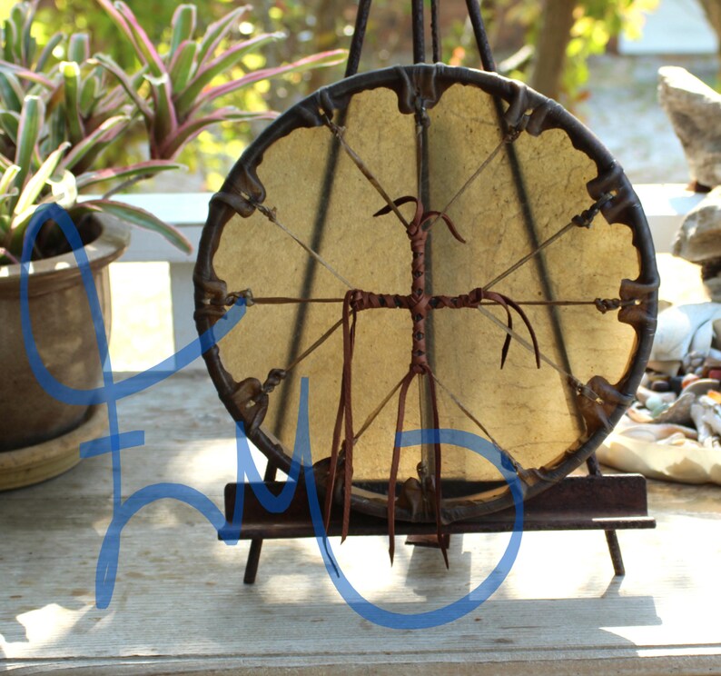 Puede incluir: Un tambor redondo, hecho a mano, con un parche de color beige claro y un marco de madera marr&oacute;n oscuro. El tambor est&aacute; sostenido por un peque&ntilde;o caballete marr&oacute;n oscuro. El dise&ntilde;o incluye un elemento en forma de cruz en el centro y detalles de cuero.