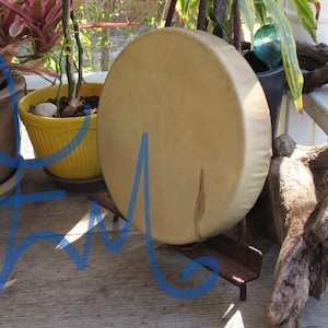 May include: A circular frame drum with a light tan, smooth skin head stretched over a wooden frame, resting upright on a small, dark brown wooden stand. The drum surface features natural variations in color and texture across the circular face.