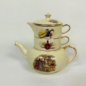 May include: Cream-colored ceramic tea set with a teapot and two stacked cups. The teapot and cups feature gold trim and colorful illustrations of musicians, wine bottles, and a bar scene. A decorative and unique tea set.