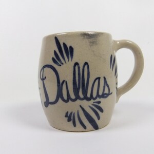 May include: A beige ceramic mug with a blue and white design. The mug has the word "Dallas" written on it in a stylized font.