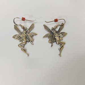 May include: A pair of silver-toned fairy earrings. Each earring features a detailed fairy figure with outstretched wings. The earrings have a hook closure with a small red bead. The background is plain white.