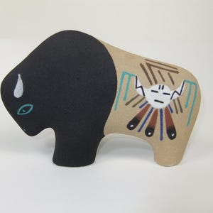 May include: A ceramic bison figurine featuring a matte black front section and a tan rear section. The tan area is decorated with a painted white mask-like face, brown geometric lines, and teal accents. A small white horn and teal eye are visible.