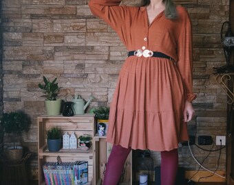 May include: A woman wearing a rust-colored dress with a black belt and a yellow beret. She is wearing red tights and red ankle boots. She is standing in front of a stone wall with a wooden shelf behind her.