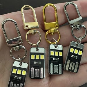 May include: A collection of keychains featuring small, black USB-shaped lights with three yellow LED lights. The keychains have silver and gold-colored clasps.