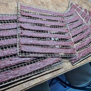 May include: Multiple metal drying racks with strips of dried beef jerky on them.