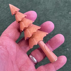 May include: A small, wooden Christmas tree figurine. The tree is light brown with a conical shape and tiered branches. The base is a light brown cylinder with a gold-colored band at the bottom. The figurine is held in a person's hand.
